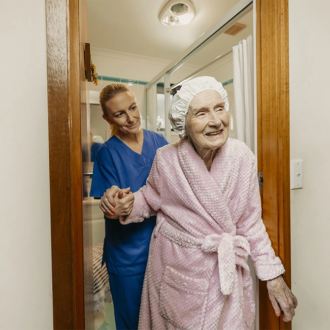 Elderly woman in pink bathrobe assisted by a young woman in blue scrubs.