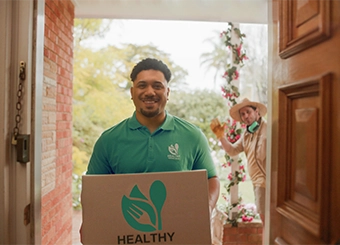 Food delivery man with package standing at the front door, with a gardener waving behind him