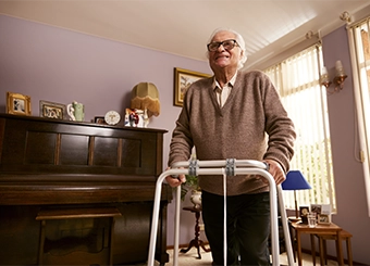 Elderly man smiling using his walking frame standing up in his living room.