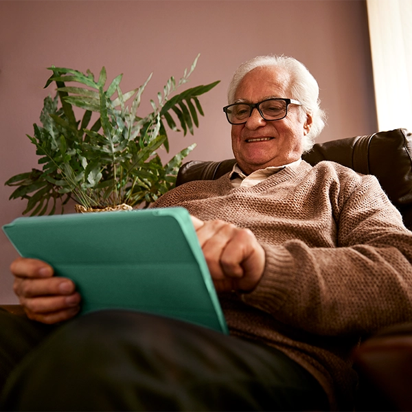 An elderly person in a brown sweater holding a teal tablet, seated in a dark brown recliner with a potted fern plant nearby.