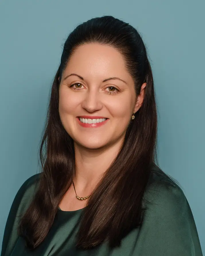 Woman seated against a teal background, wearing a dark green blouse. She has dark brown hair and is smiling