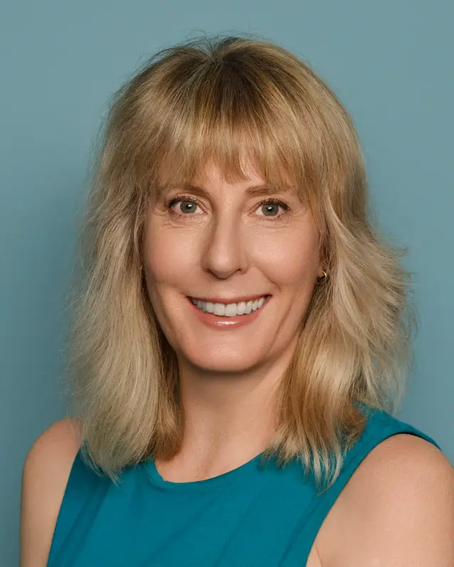 Woman in a teal dress seated on a chair against a teal background. She has blonde hair and is smiling.