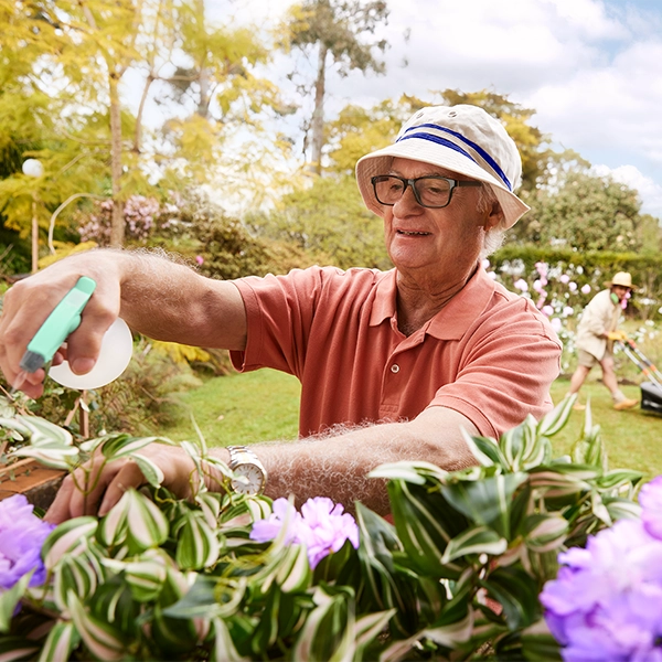 An older person in a sun hat sprays plants in a garden; another person mows the lawn in the background.