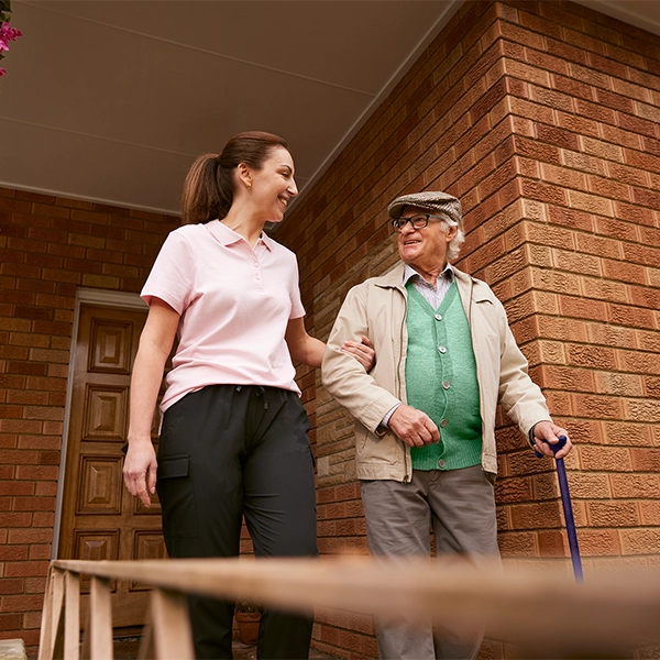 A woman helps an elderly man walk down a ramp outside a brick house.