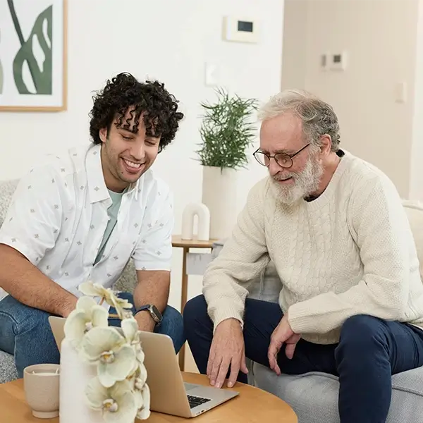 A younger man with dark, curly hair and a beard, wearing a light-colored, short-sleeved button-up shirt over a t-shirt, sits next to an older man with white hair and a beard, wearing glasses and a cream-colored sweater. The younger man has his arm around the older man's shoulders, and both are smiling and looking at a silver laptop on a wooden coffee table in front of them. There's a light-colored mug on the table. They are seated on a gray couch with light blue pillows in a well-lit room with a framed picture on the wall behind them.