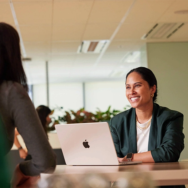 A smiling woman in a dark green blazer and white top is seated at a table with an open silver laptop in front of her. She is looking to her left at another person, whose back is to the camera. The setting appears to be a modern office with large windows and indoor plants visible in the background.