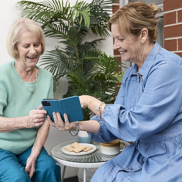 The image shows two older women sitting on a small patio or balcony, looking at a mobile phone together and smiling. The woman on the left is wearing a light teal top and dark teal or turquoise pants, and she is seated in a light-colored, woven chair. The woman on the right is wearing a light blue denim-look dress with ruffles, and she is holding the phone in a teal or dark blue case, pointing at the screen.